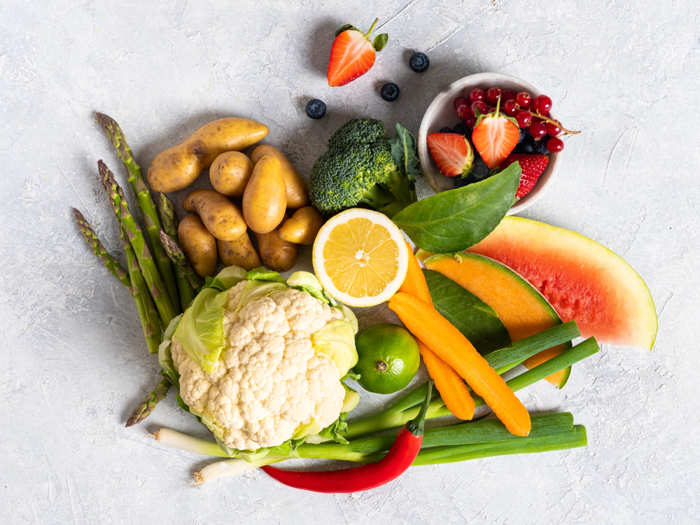 Et utvalg av frukt og grønnsaker, inkludert sitron, jordbær, vannmelon, agurk, paprika, brokkoli, blomkål, asparges, og poteter, lagt ut på en lys bakgrunn.
