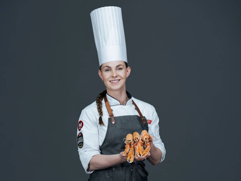 En person kledd som kokk, iført hvit kokkehatt og grå forkle, holder to kaker med sjokoladetrekk og dekorasjoner, mot en mørk grå bakgrunn.