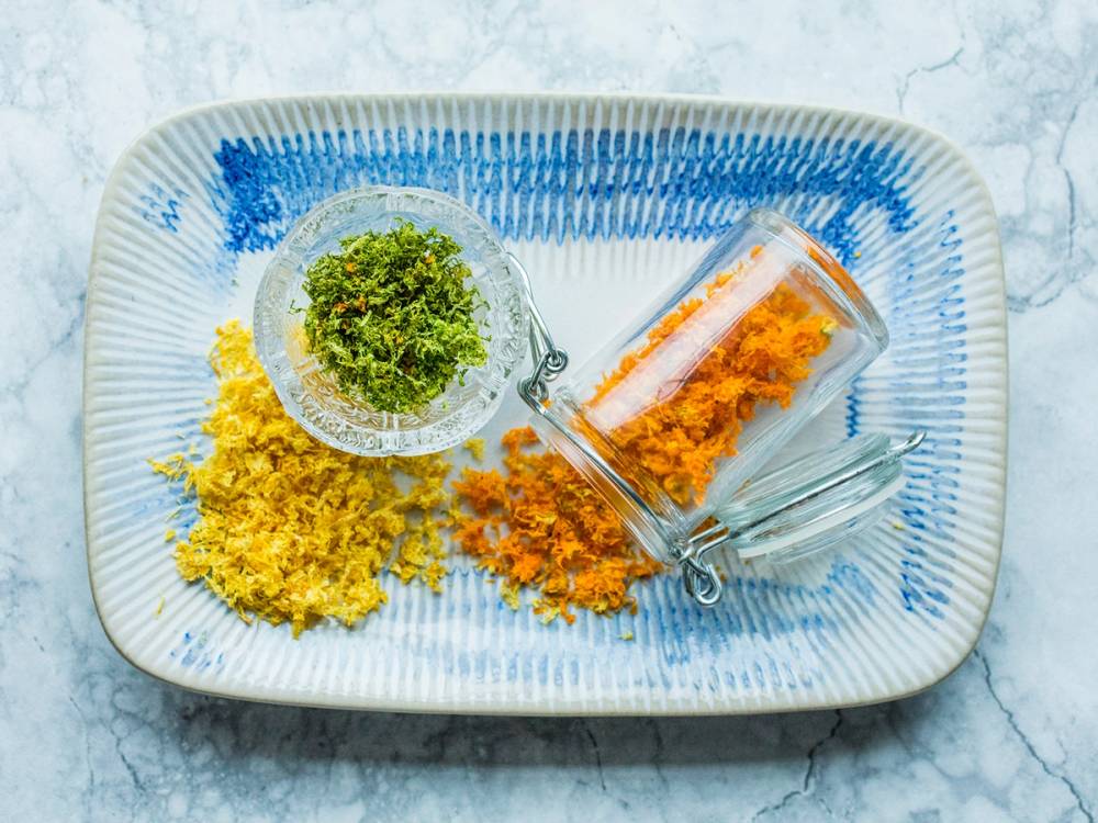 Tørkede sitrusskall i ulike farger på en hvit og blå serveringsfat med en liten skål med grønt og en glassbeholder med skallet i, på en lys steinbakgrunn.