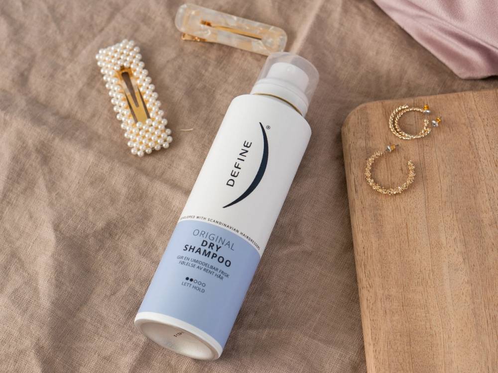 En flaske med DevaNe Original Dry Shampoo ligger på et bord sammen med hårklemmer og et par øreringer.