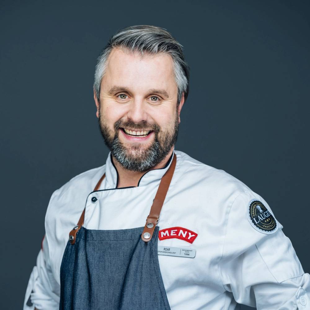 En mann i kokkeuniform med navnemerke og forkle mot en mørk bakgrunn.