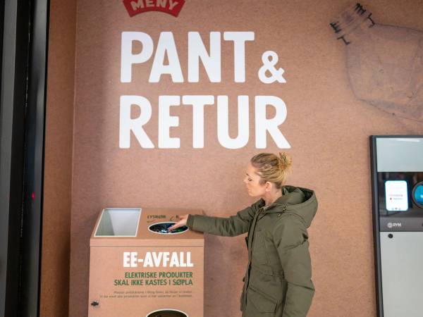 En person kaster plastemballasje i en miljøstasjon for EE-avfall, med en rosa vegg og en digital skjerm i bakgrunnen.