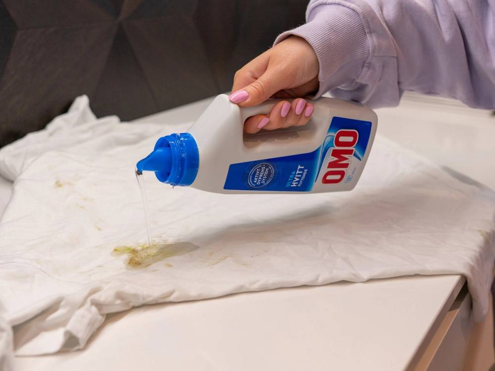 Person sprayer klut med Omo vaskemiddel på et hvitt underlag.