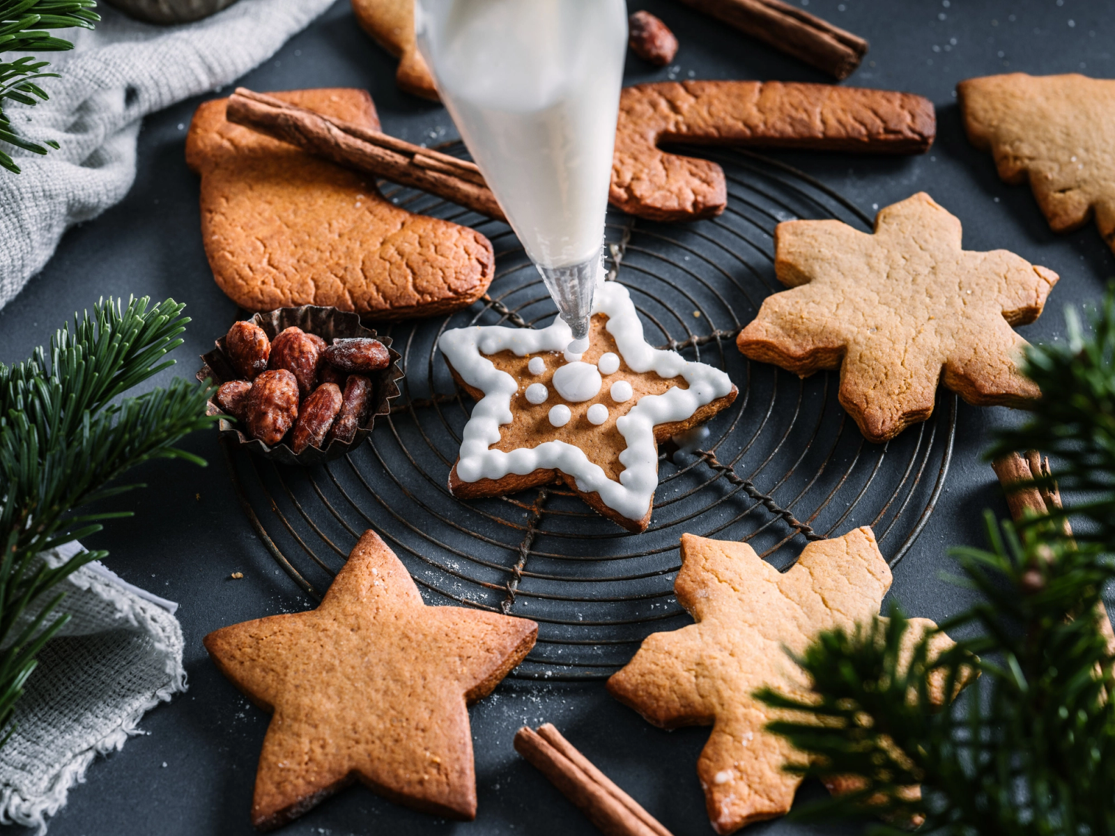 Pepperkaker i ulike former ligger på en svart rist med pynt og kanelstenger rundt, mens en sprøyte fyller en pepperkake med hvit glasur.