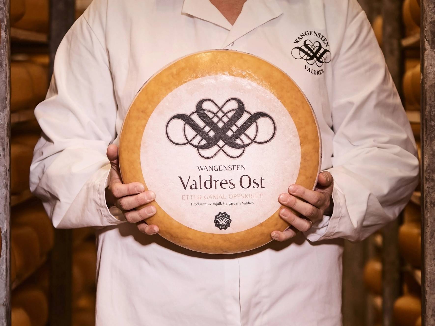 Person i hvit kokkeforkle holder en stor ost med etiketten Valdres Ost fra Wangenstein.