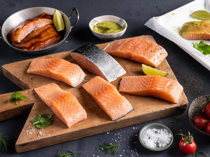 Ulike laksefileter og lakseporsjoner på et skjærebrett med sitronbåter og urter, klar for tilberedning, på en mørk bakgrunn.