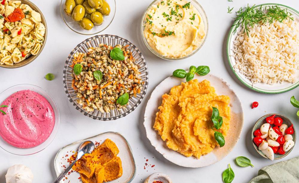 Forskellige typer saus og tilbehør på små tallerkener og skåler, inkludert hummus, ris, og grønnsaker.