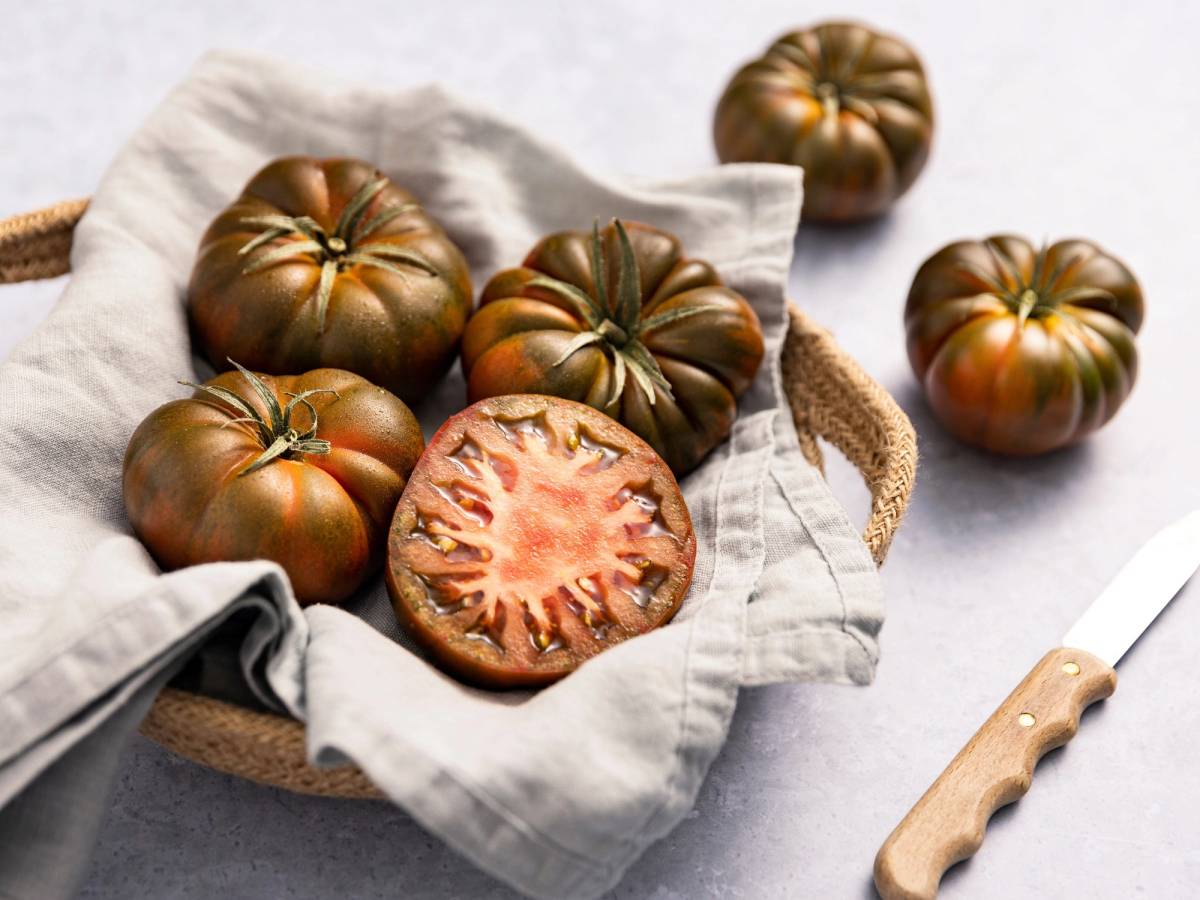 Tomater med mørkebrune og grønne farger ligger i en kurv med en skåret tomat som viser innsiden, på en hvit klut.