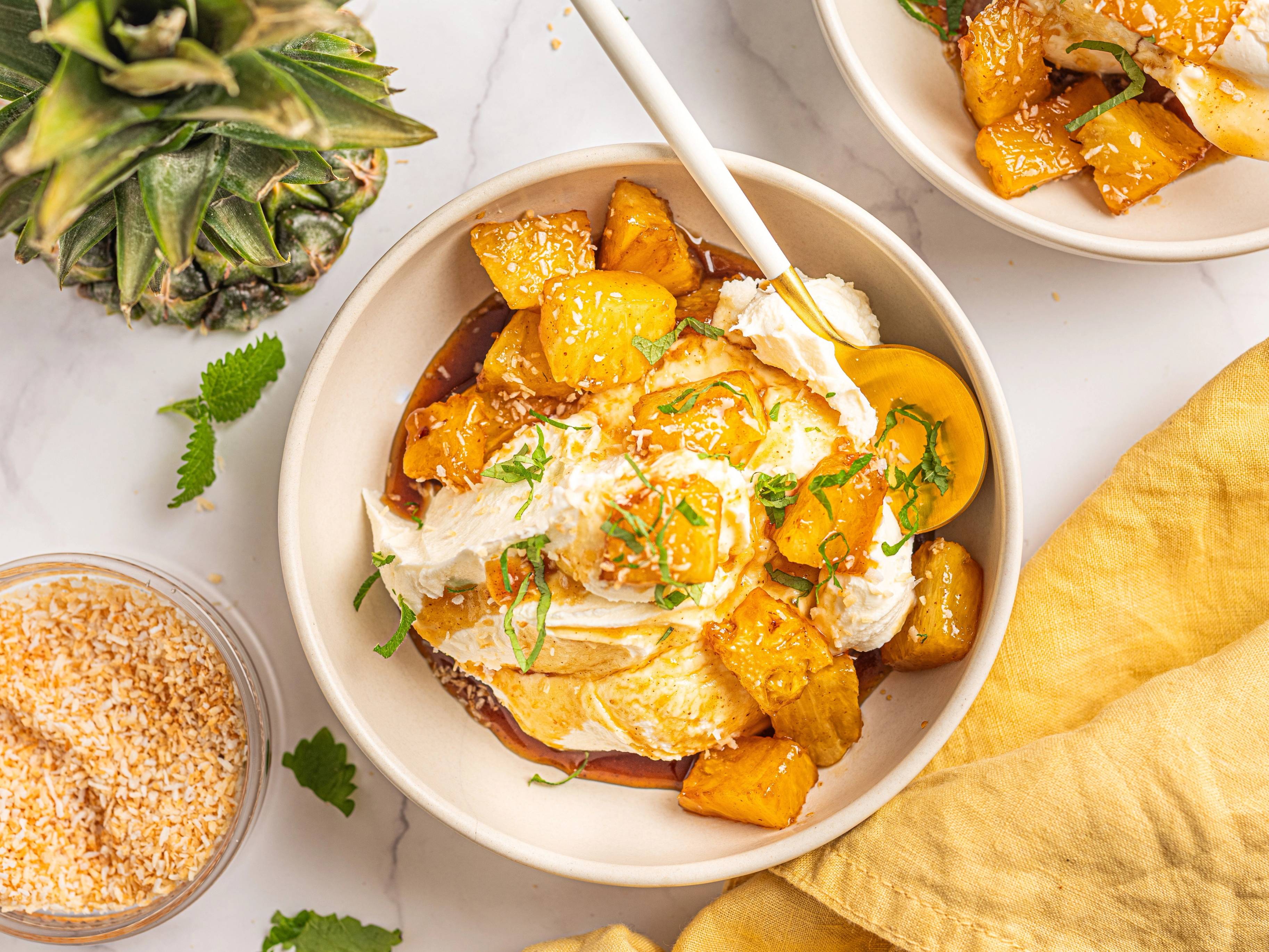 En skål med mascarponekrem og glasert ananas, pyntet med grønne urter, omgitt av andre småretter og frukt på et bord.