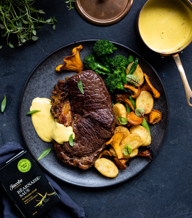 Stekt biff med ovnsstekte poteter, brokkoli og sopp på en svart tallerken, med saus og urter ved siden av.