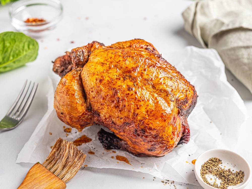 Nygrillet kylling med sprø skorpe ligger på hvitt papir, omgitt av krydder og grønnsaker.