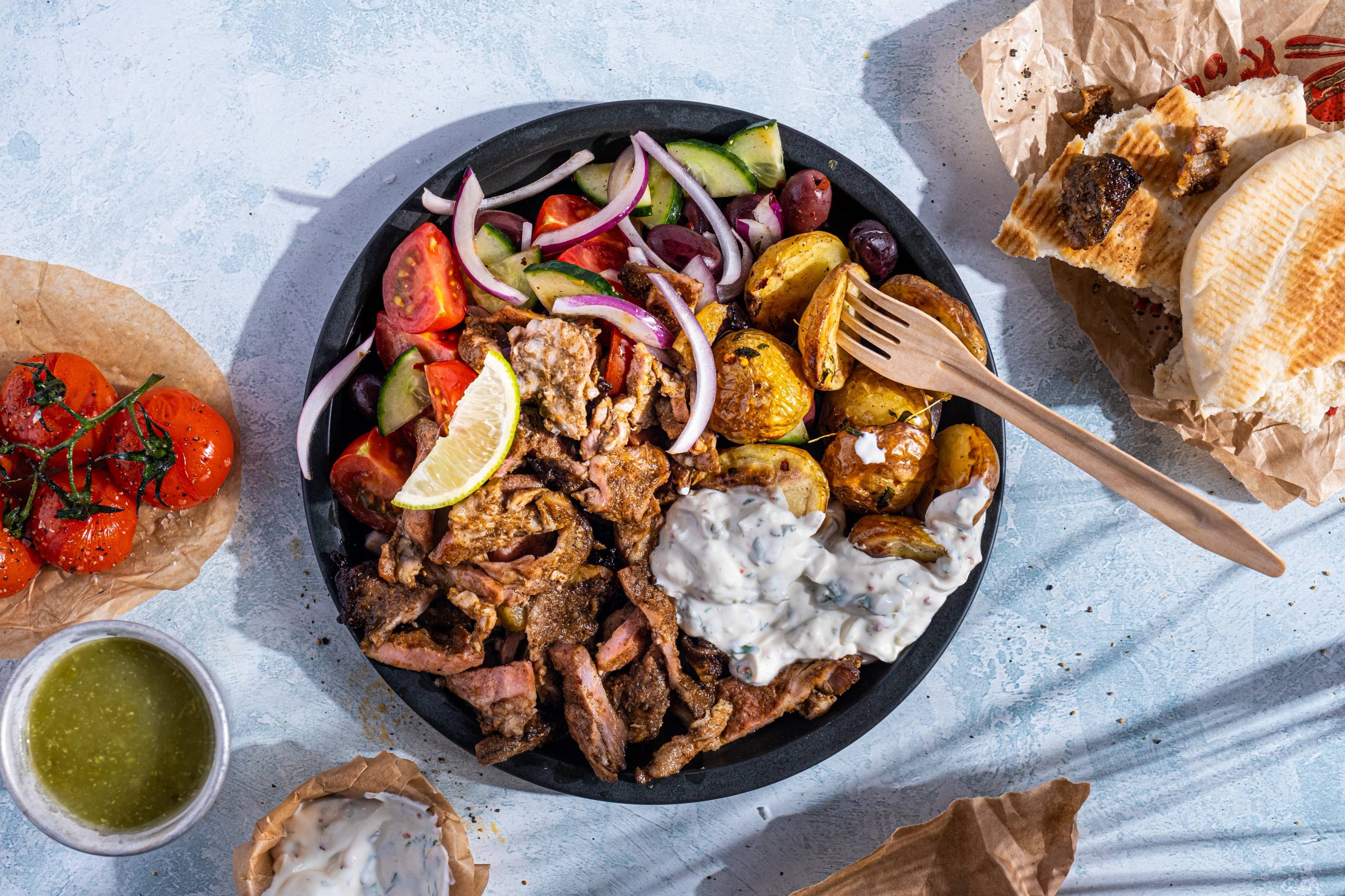 En tallerken med grillet svinekjøtt, ovnsbakte poteter, tomatsalat og en skål med hvit saus, omgitt av brød og en liten skål med grønn saus.