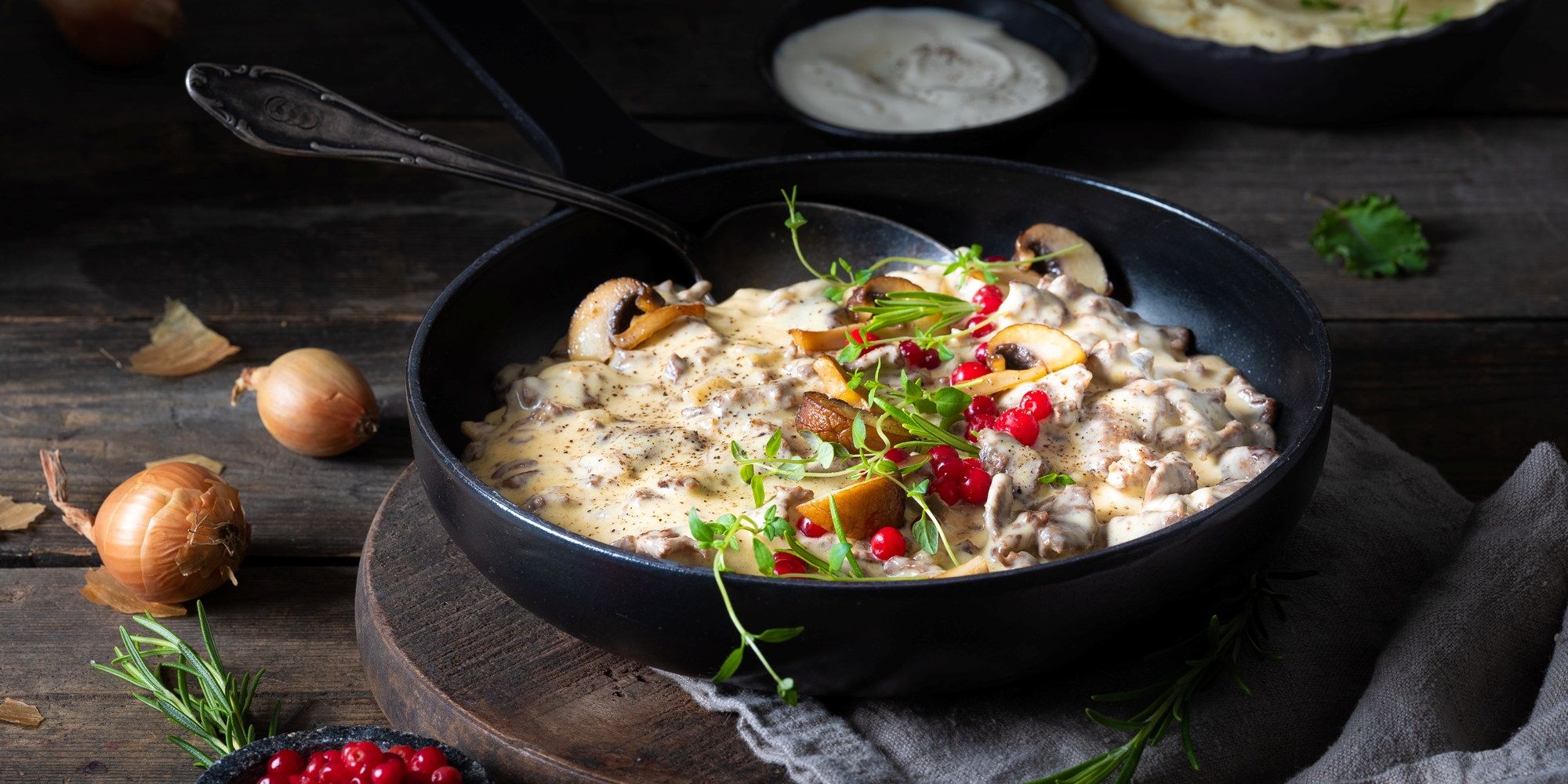 En skål med kremet sopprett servert med urter og granateplefrø, omgitt av sopp og urter på en mørk treplate.