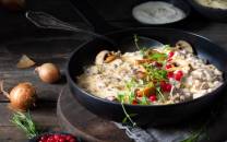 En skål med kremet sopprett servert med urter og granateplefrø, omgitt av sopp og urter på en mørk treplate.