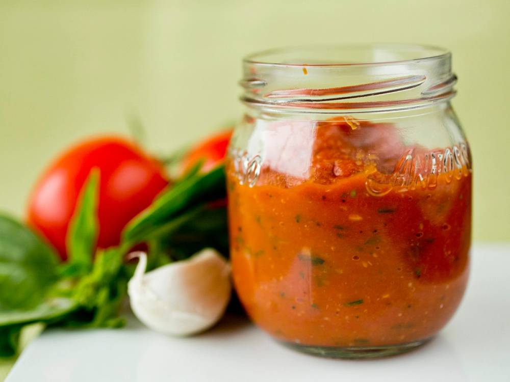 Hjemmelaget pasta med tomatsaus i et glass, omgitt av tomater, hvitløk og ferske urter.