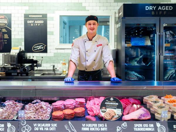 En person i kokkeforkle på en kjøtt- og pølsebod i en butikk eller marked, med kjøttprodukter foran seg.