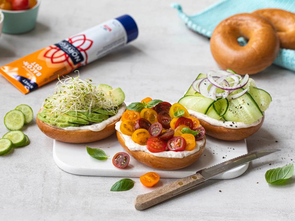 Bagels med smøreost, agurk, tomater og løk på en hvit tallerken, med et par donuts i bakgrunnen.
