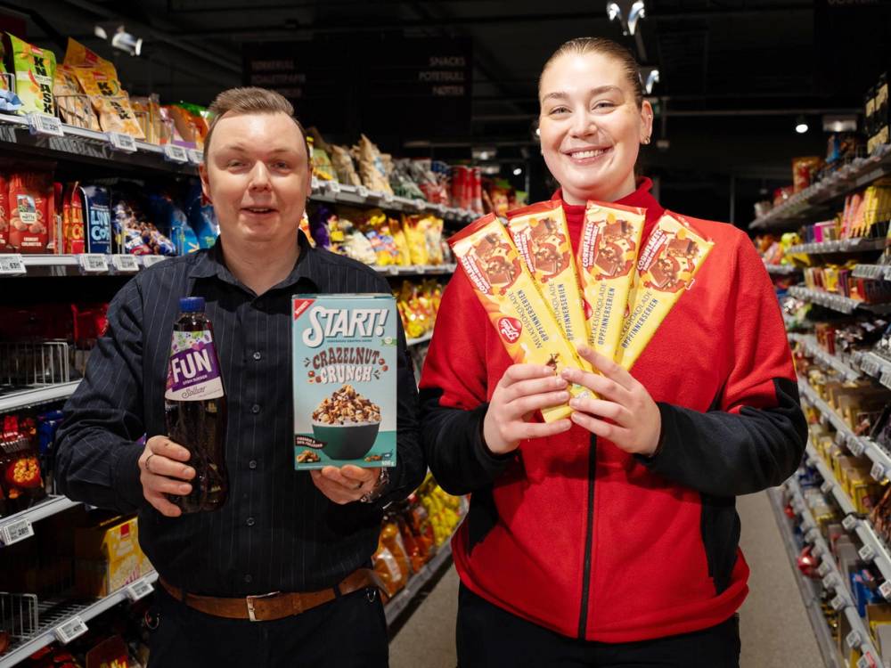 To personer står i en dagligvarebutikk, den ene holder en flaske med brus og den andre viser frem flere gule og røde pakker, sannsynligvis snacks eller godteri.