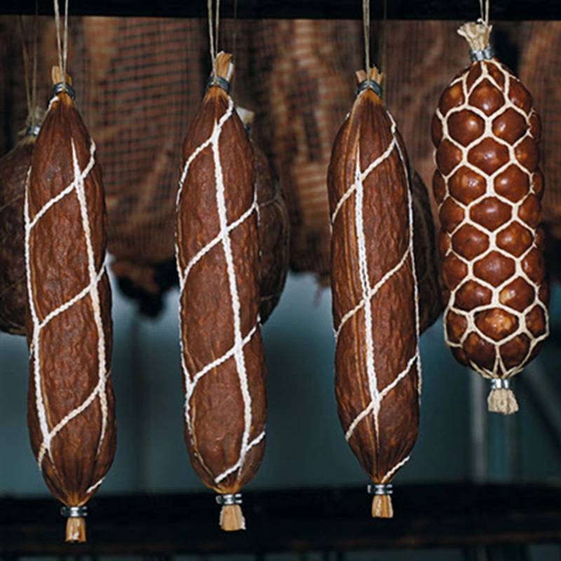 Fire spekepølser henger til modning med en stor spekepølse i bakgrunnen som har et honeycomb-mønster av hvite nettingbånd.