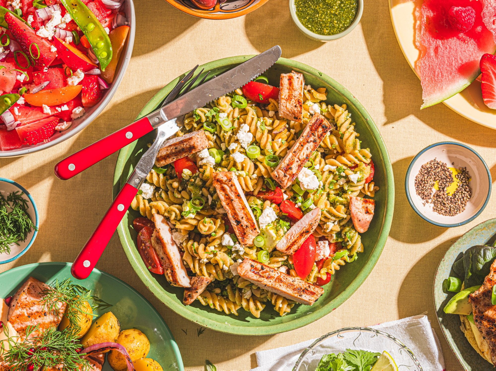 En stor grønn bolle med pastasalat som inneholder pasta, cherrytomater, agurk og skiver av kjøtt, omgitt av flere små boller med ulike salater og dressinger.