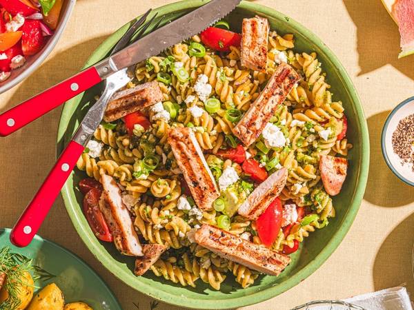 En stor grønn bolle med pastasalat som inneholder pasta, cherrytomater, agurk og skiver av kjøtt, omgitt av flere små boller med ulike salater og dressinger.