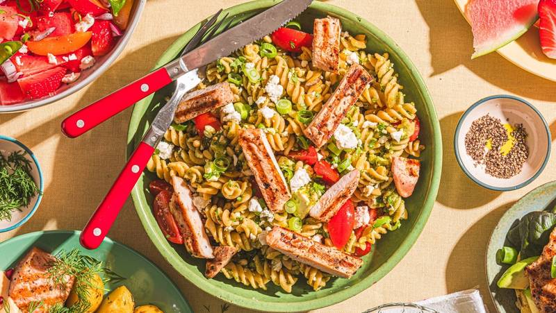 En stor grønn bolle med pastasalat som inneholder pasta, cherrytomater, agurk og skiver av kjøtt, omgitt av flere små boller med ulike salater og dressinger.