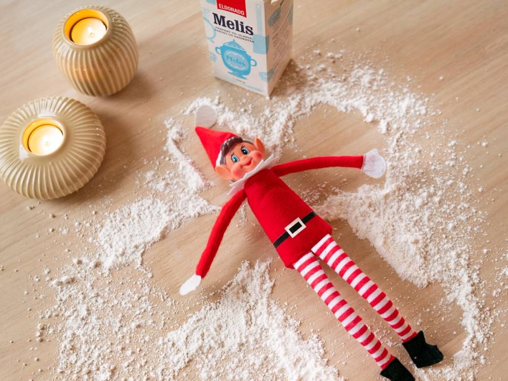 En jule-nisse i rød drakt og stripete bukser ligger på et dekket bord med mel og to lyslykter rundt seg.
