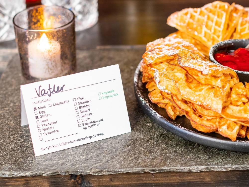 En tallerken med vafler og en liten skål med bær, plassert på en steinplate med et lysende stearinlys ved siden av, og en matliste i bakgrunnen.