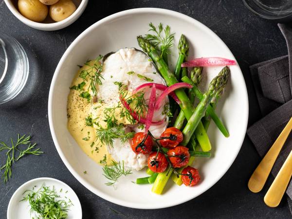 Skrei med hollandaise-saus servert med grønne asparges, cherrytomater og rødløk på en hvit tallerken.