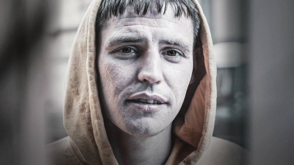 Person med hettegenser som har ansiktet dekket av en grå skjerm, står i et rom med bokhyller i bakgrunnen.