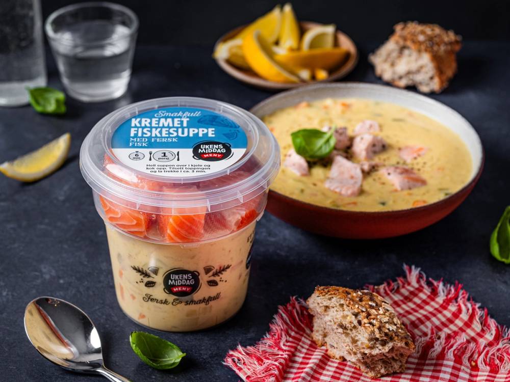 Kremet fiskesuppe i en plastkopp med skje ved siden av, omgitt av sitronbåter og brød på en mørk bakgrunn