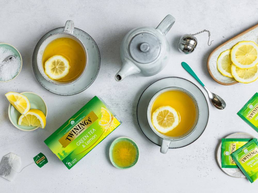 To kopper med te med sitron, en tekanne, og pakker med Twinings te på et hvitt bord.