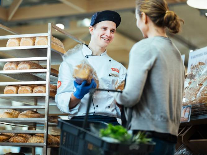En ansatt i en bakeributikk hjelper en kunde med å velge sunnere brødvalg, mens de står foran hyller med ulike typer brød.