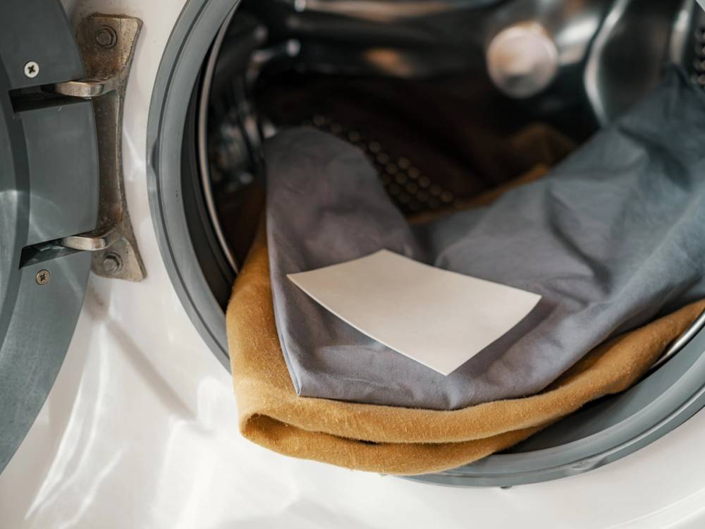 En person som sitter i en bil med en hvit papirlapp på fanget, iført grå bukser og en brun jakke, sett fra passasjersiden.