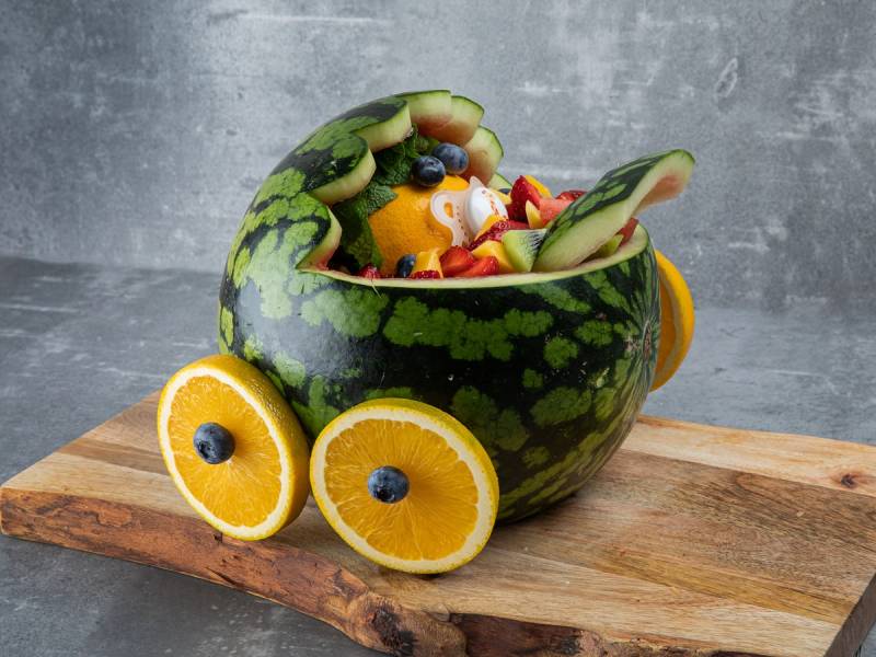 En dekorativ fruktsalat i en vannmelonskål med hjul laget av sitron- og blåbærskiver, plassert på et treplate.