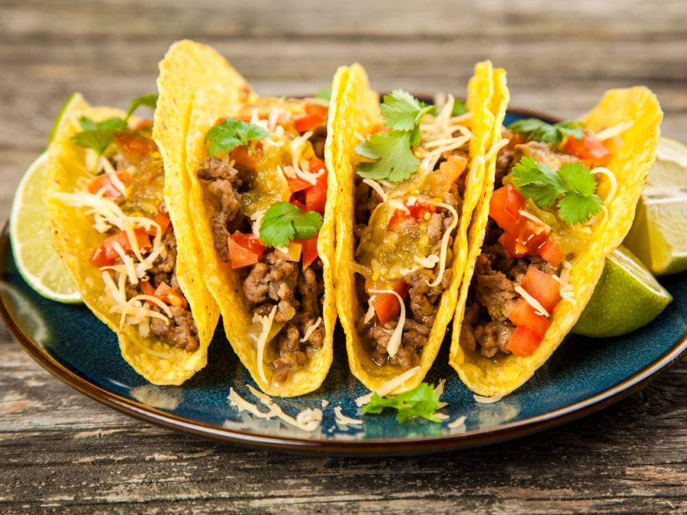 Fire tacos fylt med kjøtt, grønnsaker og ost, servert med limebåter på en blå tallerken.