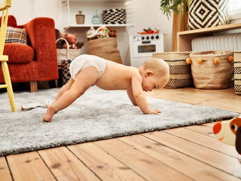 Baby som kryper på gulvet i stue med sofa og lekekasser i bakgrunnen.