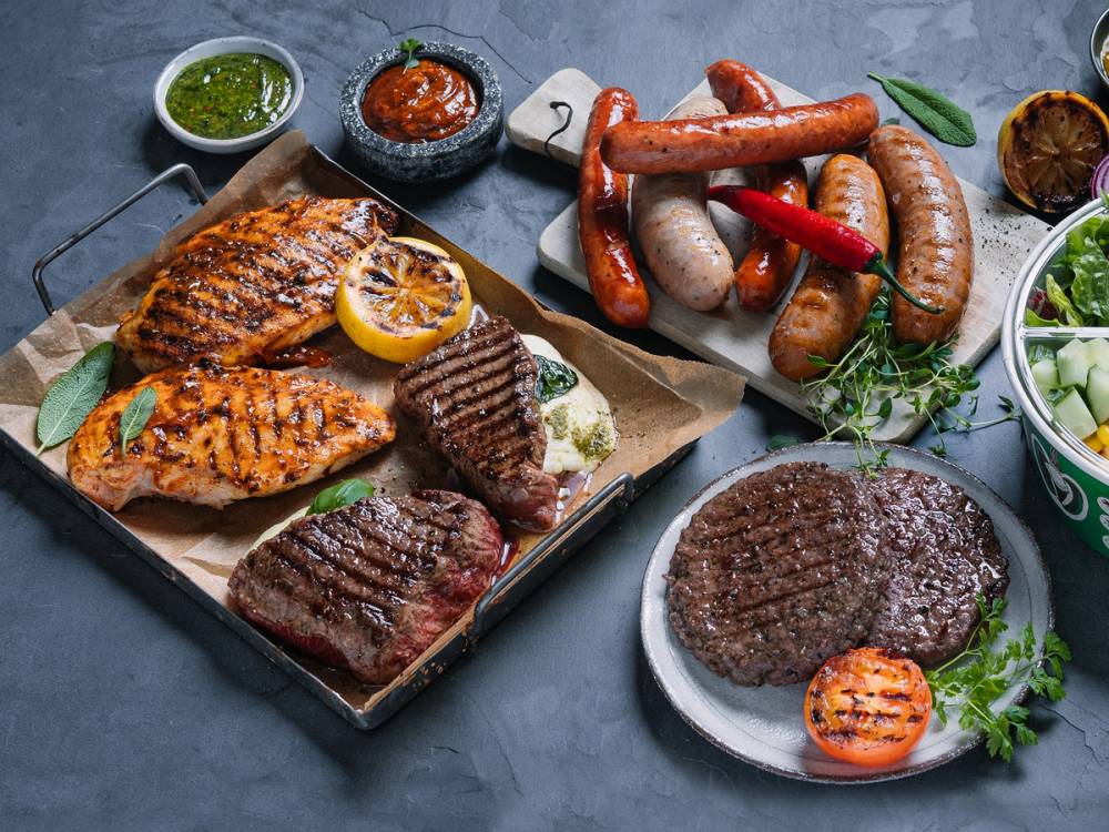 Grillmat med ulike kjøttstykker og pølser på et bord, omgitt av sauser og grønnsaker.