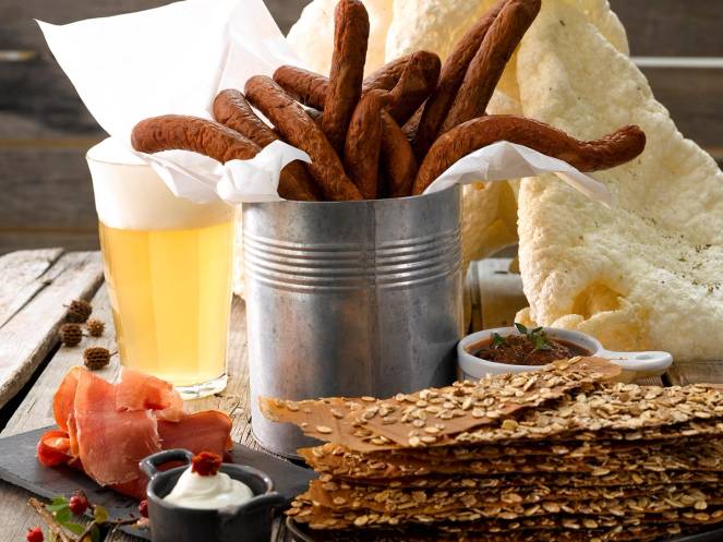 En tallerken med sprø kjeks, en skål med rømme, og et glass med øl eller cider, sammen med en metallbøtte fylt med pølse og potetgull på et bord.