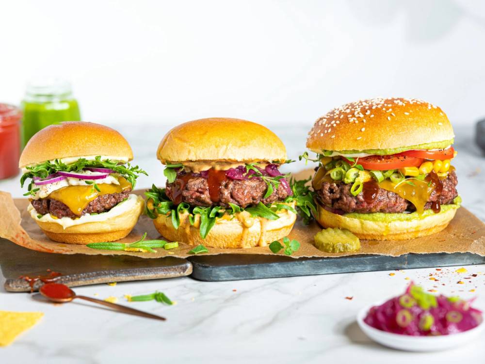 Tre burgere med ulike fyll, servert på en steinplate med tilbehør og grønnsaker i bakgrunnen