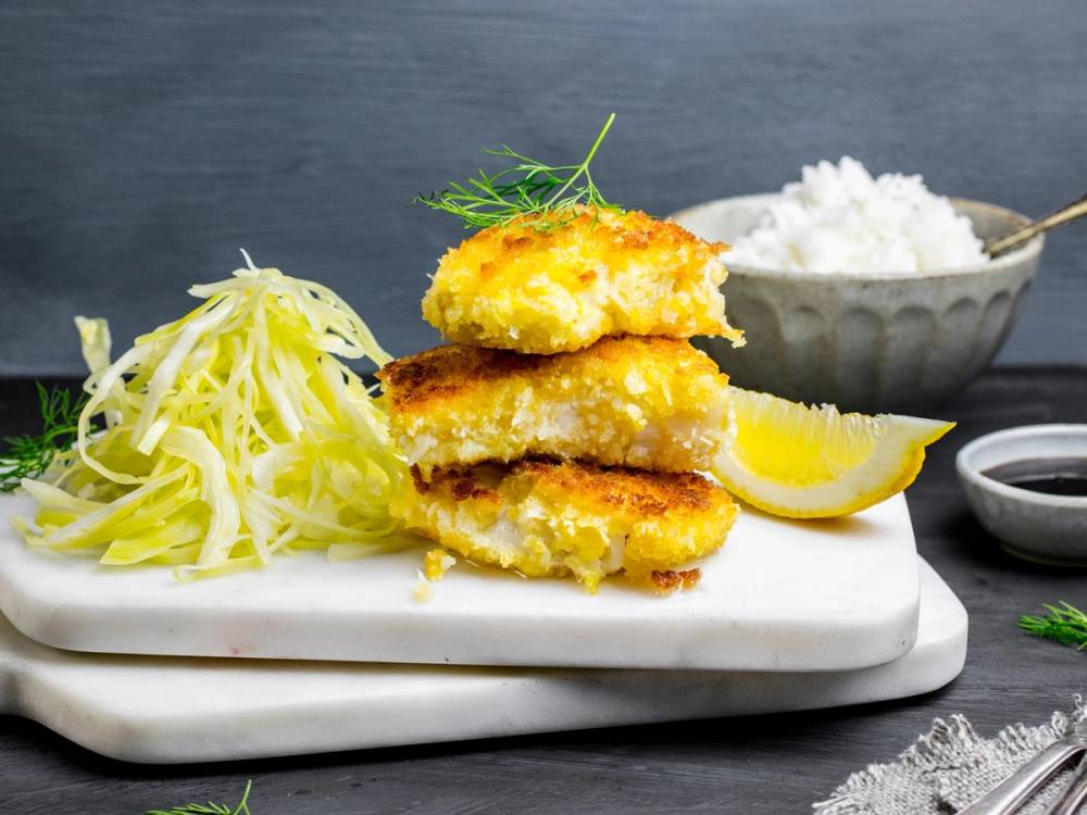 Pankopanert torsk med kålsalat, ris, sitron og en liten skål med hvit saus på en hvit tallerken.