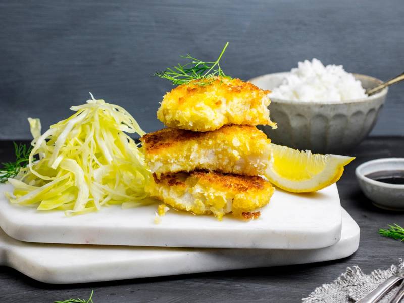 Pankopanert torsk med kålsalat, ris, sitron og en liten skål med hvit saus på en hvit tallerken.