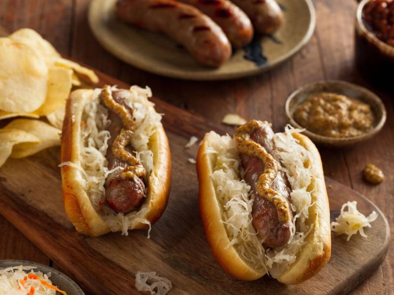 Pølser i brød med kål og sennep, typisk for oktoberfest, på et trebord med en liten skål med sennep ved siden av.