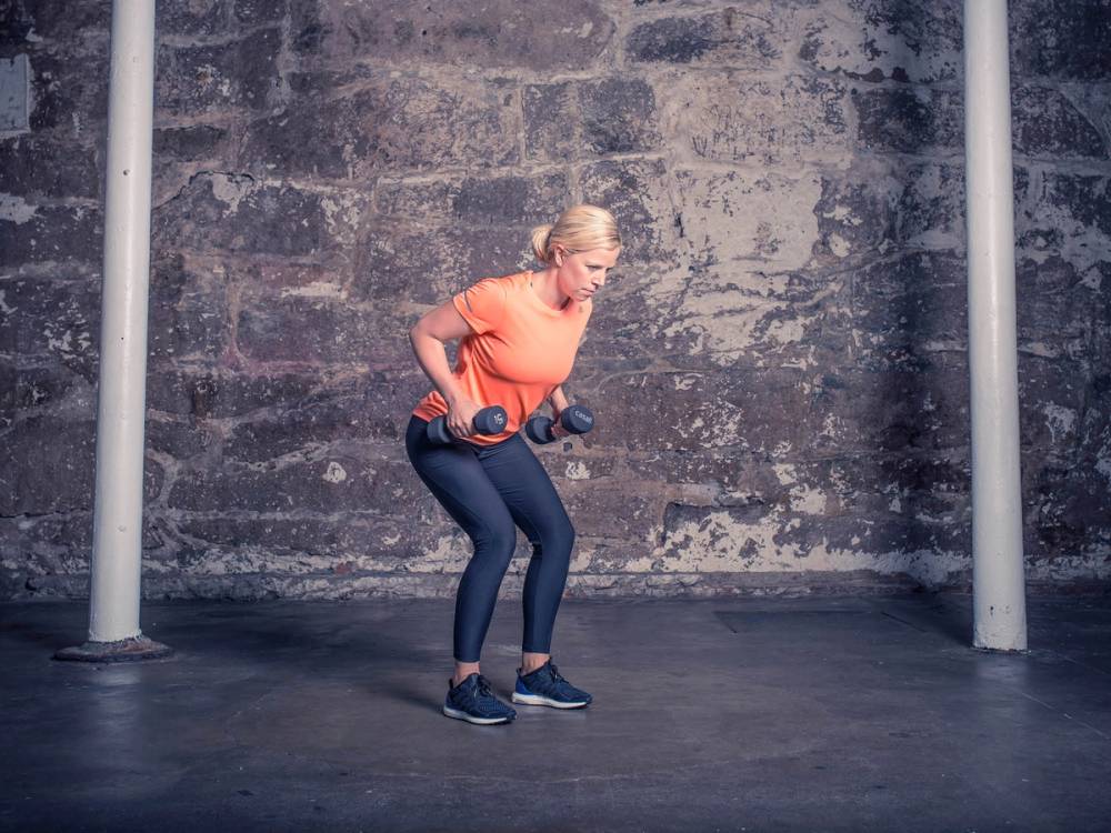 En kvinne trener med manualer i en treningssal med betongvegg i bakgrunnen.