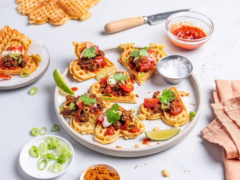 Vaffel nachos med kjøtt, salsa, rømme og lime på en hvit tallerken, omgitt av tilbehør og en kniv.