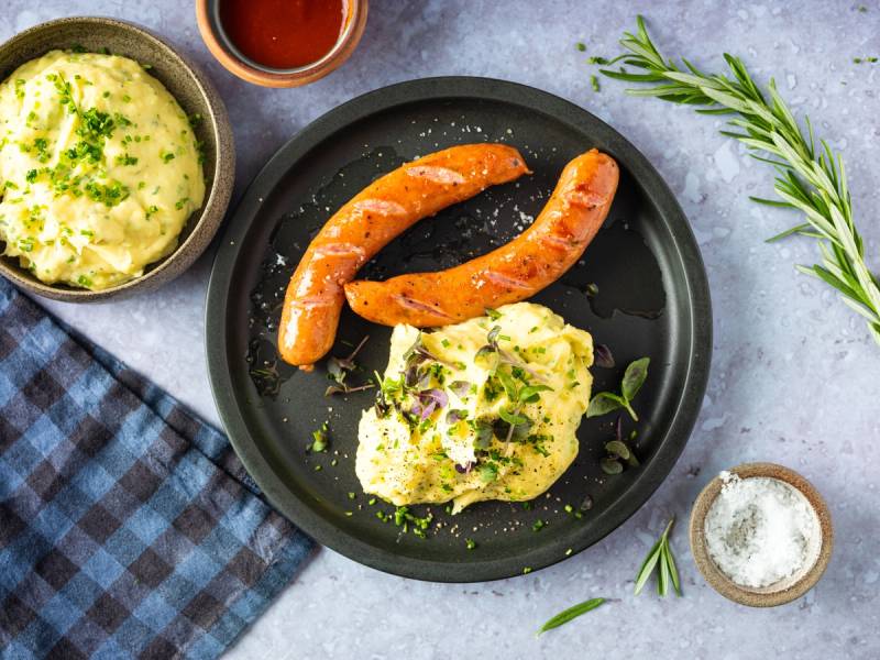 Potetmos med grønne urter, to pølser og en liten skål med salt på en svart tallerken, sett på et bord med en blått og hvitt rutete serviett og noen urter.