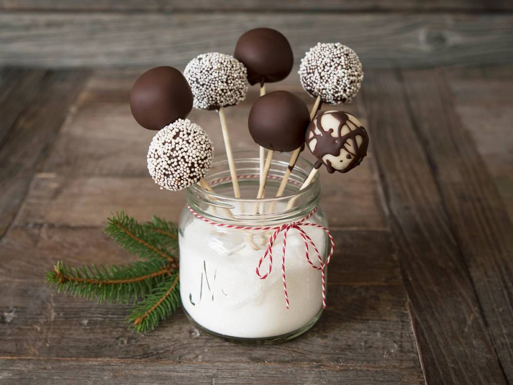 Hjemmelagde marsipanskulpturer på pinne i en glasskrukke med en liten granbar ved siden av, satt på et trebord.