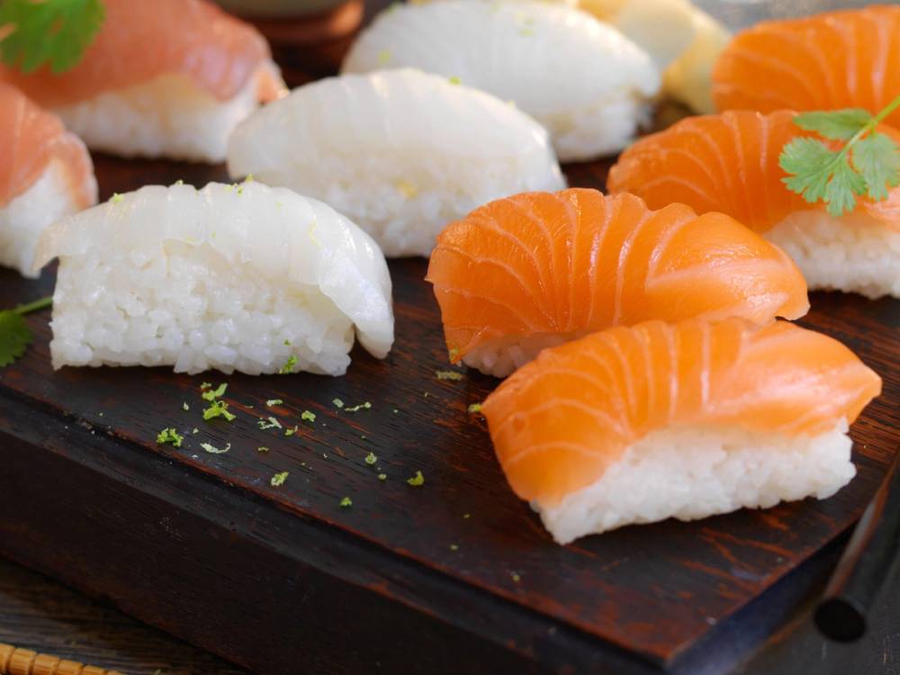 Sushi med laks og hvit fisk på en svart serveringsplate, pyntet med sitronskiver og grønn koriander.