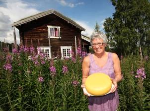 En person står i en blomstereng foran en gammel trehytte, og holder en stor gul ostekake.