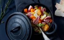 Lapskaus med kjøtt, poteter og grønnsaker i en svart gryte med lokk ved siden av, klar til servering.